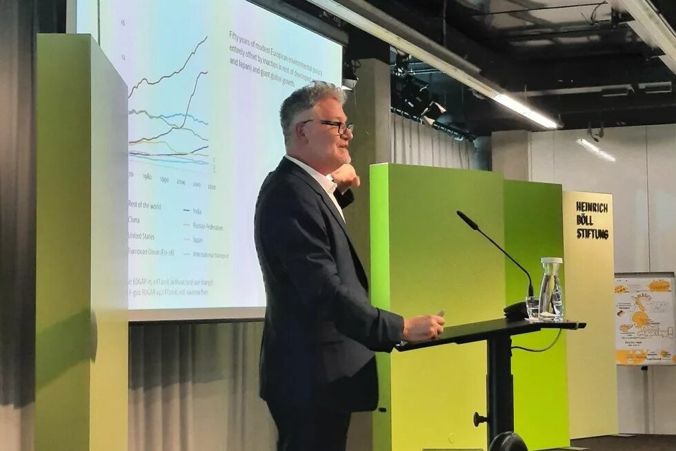 Adam Tooze A man speaks at a podium with a microphone; behind him are a graph and the Heinrich Böll Foundation logo.