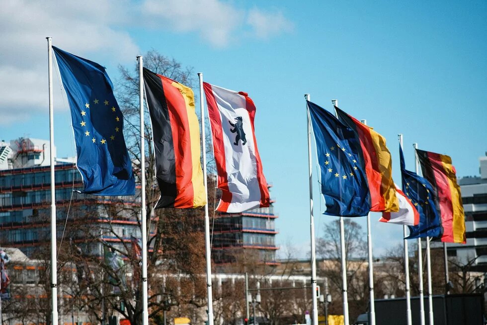 Europa-, Deutschland- und Berlin-Flaggen an Fahnenmasten wehen vor Gebäuden im Hintergrund-