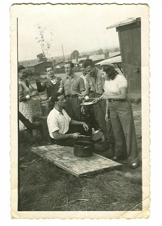 Sieben Personen stehen oder hocken im Freien bei einer Essensausgabe neben einem Holzverschlag.
