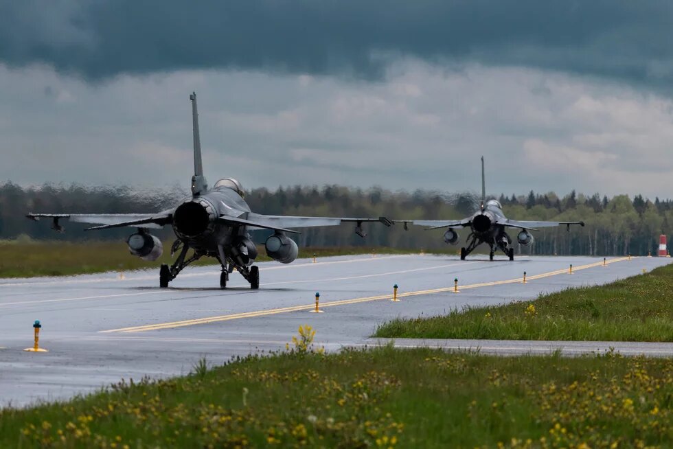 Portuguese Air Force Zwei Kampfjets rollen hintereinander auf nasser Landebahn, Wiese und Wald rechts und links, dunkle Wolken am Himmel.
