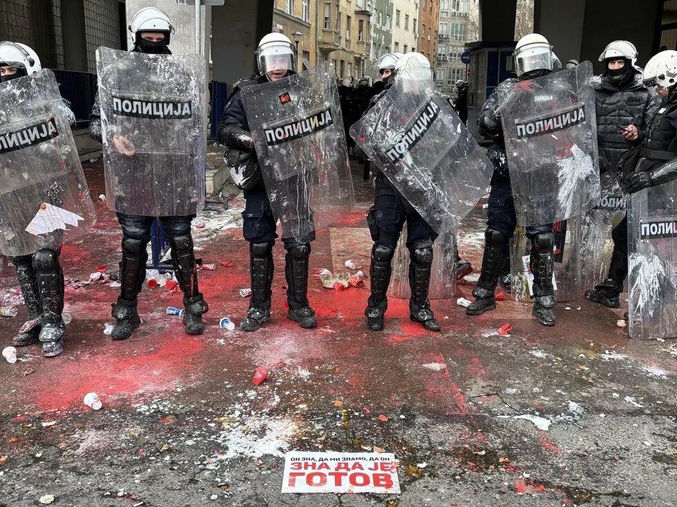 Police Officers in Novi Sad, Serbia Police officers with shields, surrounded by red powder and trash on the street. sign on the ground says in Serbian: “He knows that we know that he is finished”