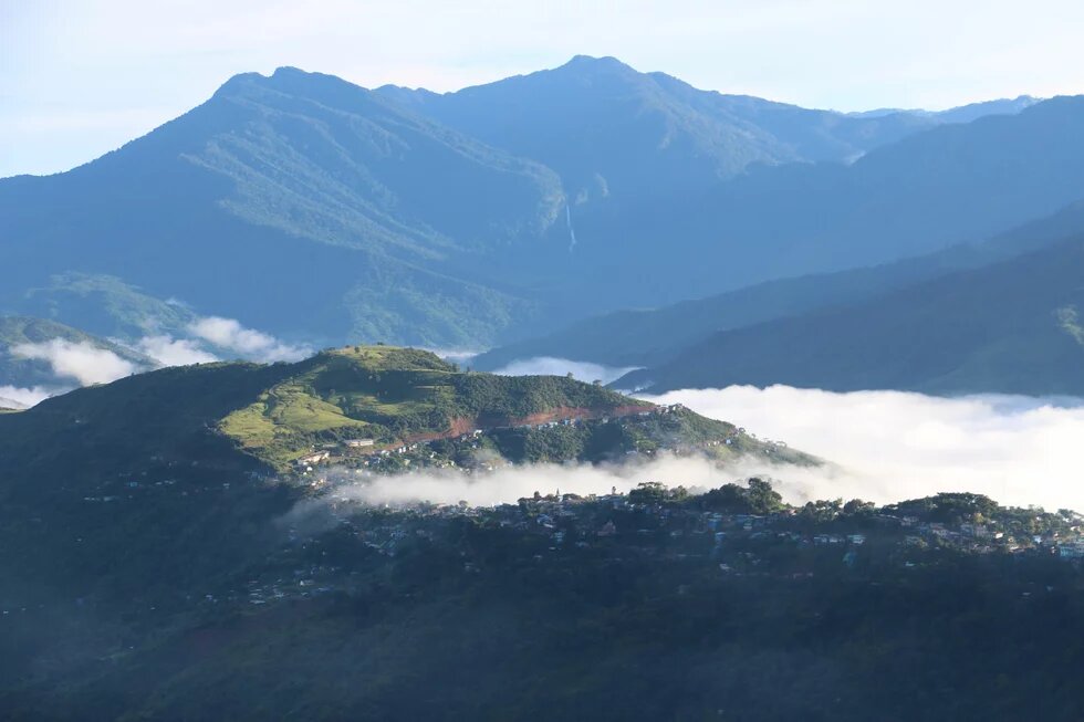 Built-up hillside in morning light, clouds drifting through the valley, tall forested mountains in the background.