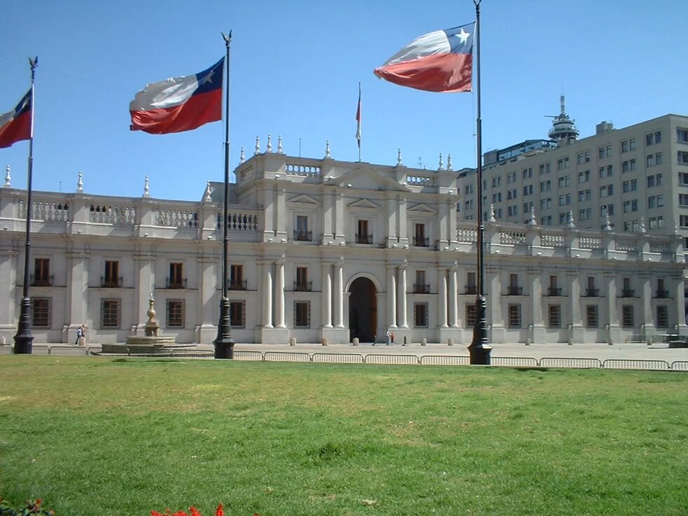 Foto des Präsidentenpalastes "La Moneda" in Santiago de Chile mit Vorplatz und Fahnenstangen mit gehisster chilenischer Flagge.
