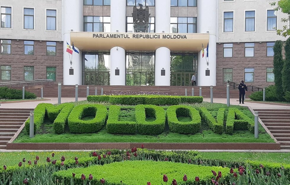 Vor dem Parlamentsgebäude der Republik Moldau steht in Buchsbaum das Wort „MOLDOVA“. Davor Blumenbeete, zwei Personen auf den Treppen.