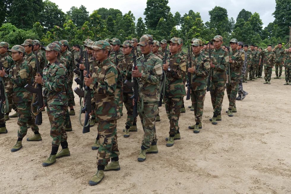 Tabayin People’s Defense Force Group of soldiers in camouflage uniforms with rifles marching on sandy ground, trees in the background.