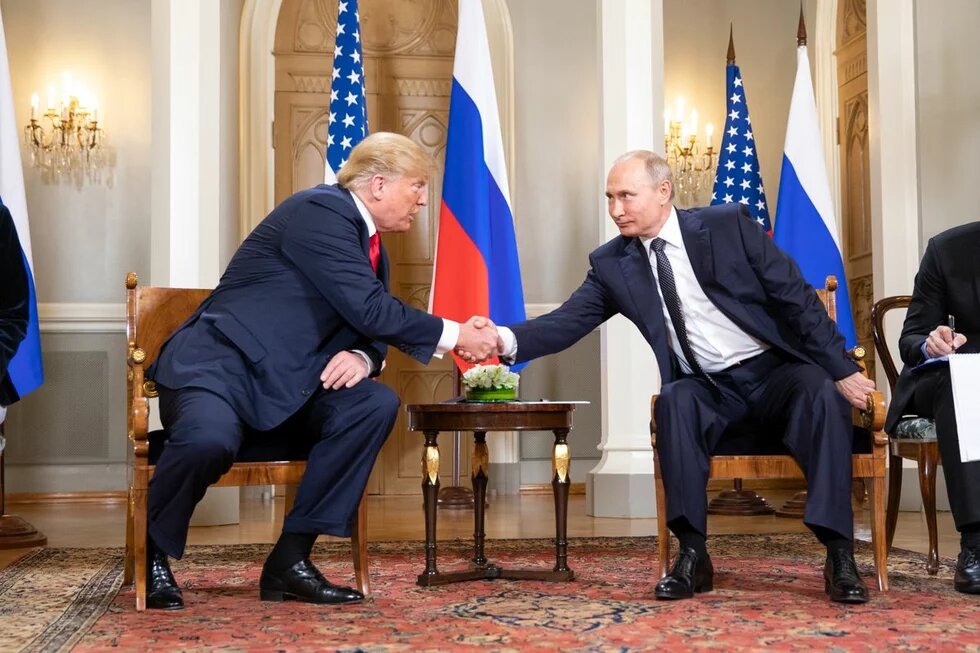 US President Donald J. Trump and President Vladimir Putin of the Russian Federation Donald Trump and Vladimir Putin sit facing each other and shake hands. A small table with flowers is between them.