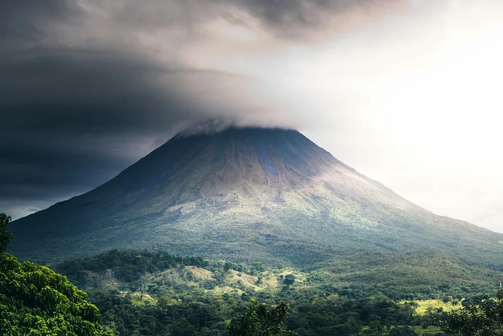 A lone volcano rises from dense vegetation, its peak covered by dark clouds.