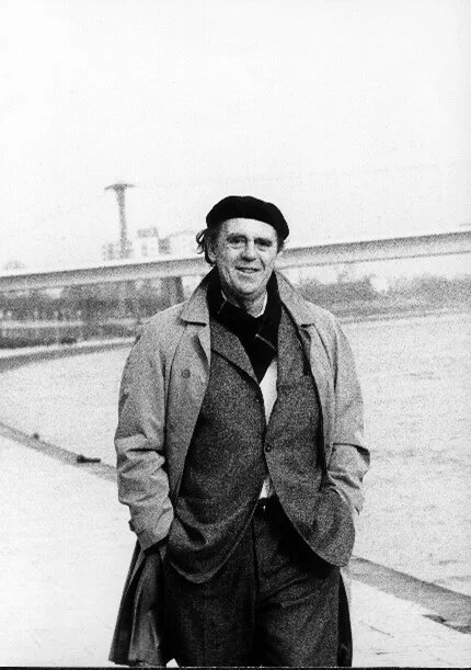 Heinrich Böll mit Mantel und Mütze geht lächelnd am Fluss Rhein entlang; im Hintergrund eine Brücke und Industrieanlagen.
