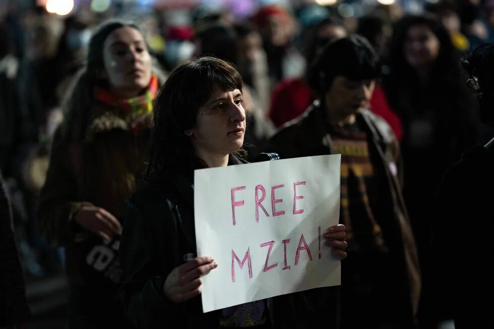 Person hält ein Schild mit der Aufschrift „FREE MZIA!“ in einer Menschenmenge bei Dunkelheit.