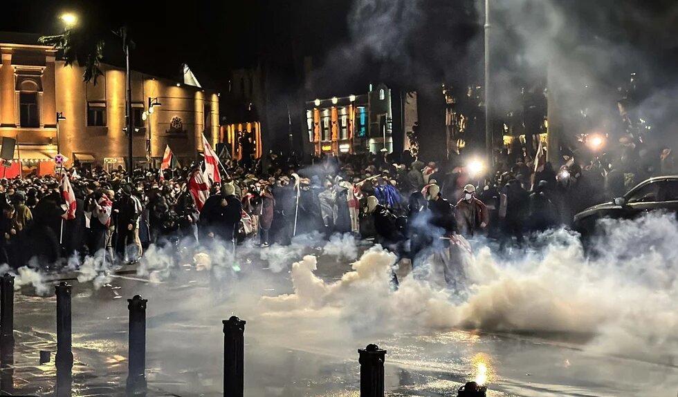 Große Menschenmenge bei Nacht, viele Flaggen, Rauch steigt auf, einige Personen tragen Masken, Lichtquellen im Hintergrund.