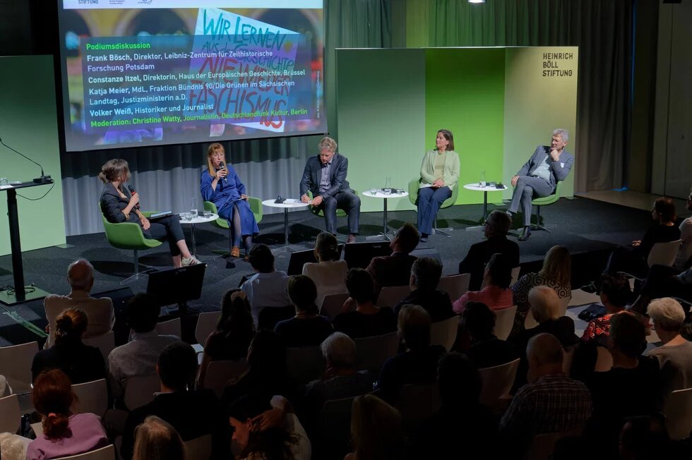 Fünf Personen auf Podium mit Mikrofonen und Notizen, Publikum sitzt davor, im Hintergrund Projektion mit Namen und Titel.