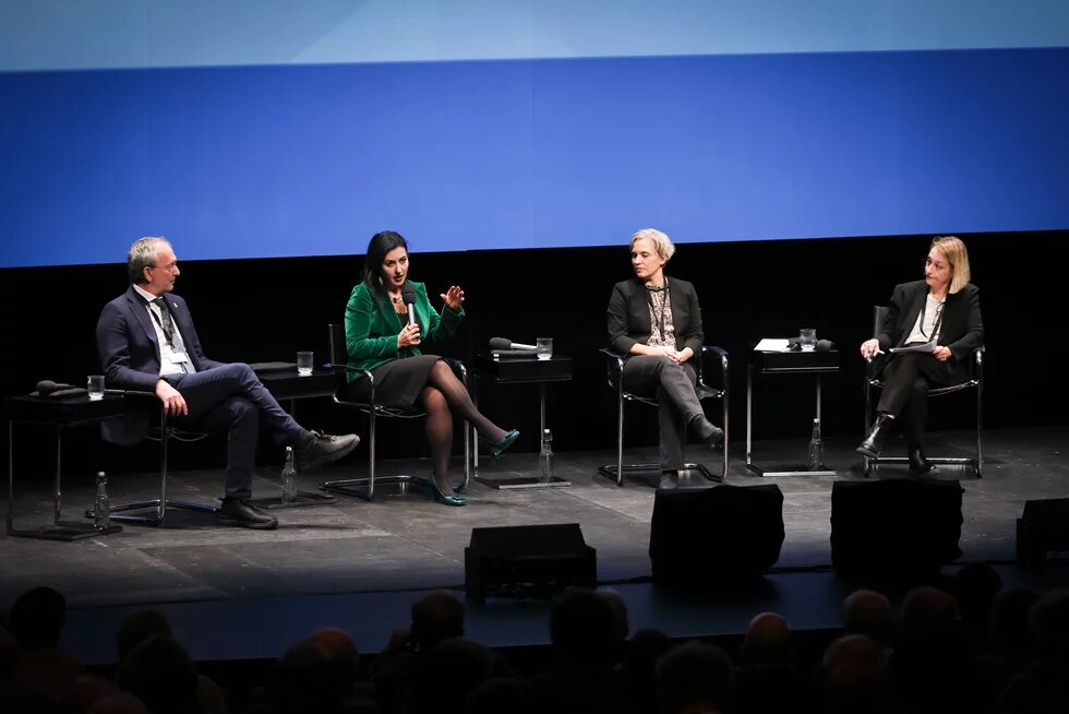 Vier Personen sitzen auf einem Podium, im Hintergrund scheint blau-weiß eine Leinwand. 
