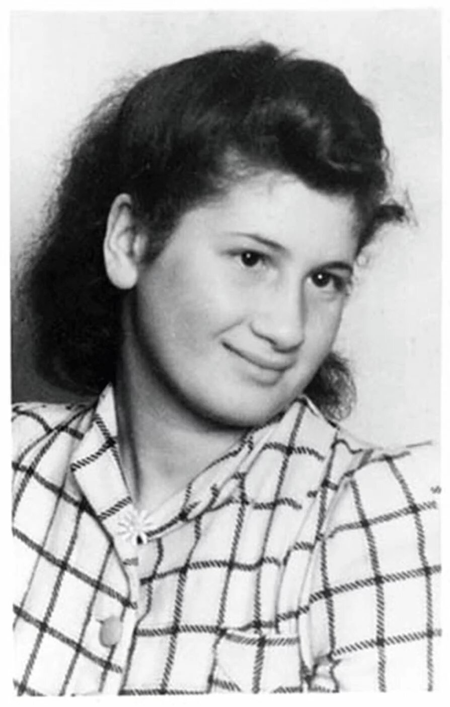 Porträtaufnahme einer Person mit kariertem Oberteil, leichtem Lächeln und seitlichem Blick, neutraler Hintergrund.