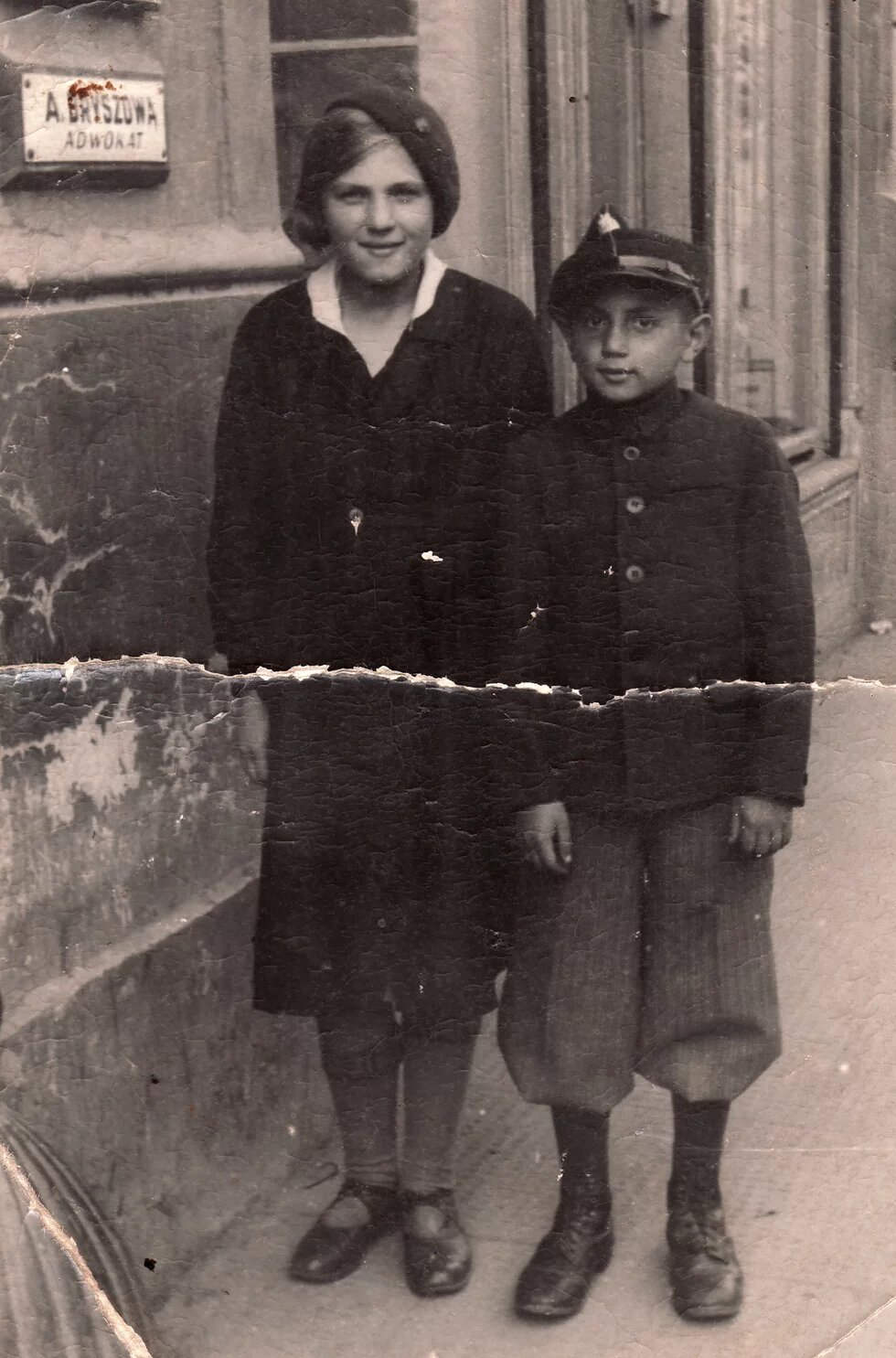 Zwei Kinder stehen auf einem Gehweg vor einem Gebäude mit einem Schild mit der Aufschrift „A. Griszczuk – Adwokat“.