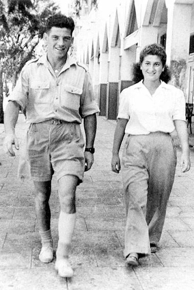 Zwei Personen gehen auf einem Gehweg nebeneinander, im Hintergrund eine Häuserreihe mit Arkaden.