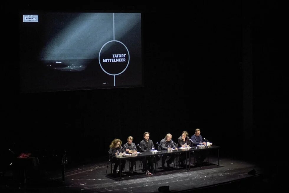 Mehrere Personen sitzen auf einer Bühne mit einer Leinwand im Hintergrund.