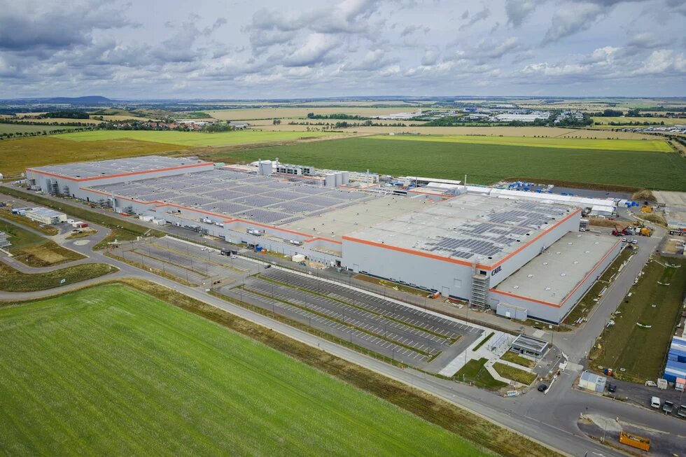 Large industrial building with solar panels on the roof, surrounded by fields, roads, and mostly empty parking lots.