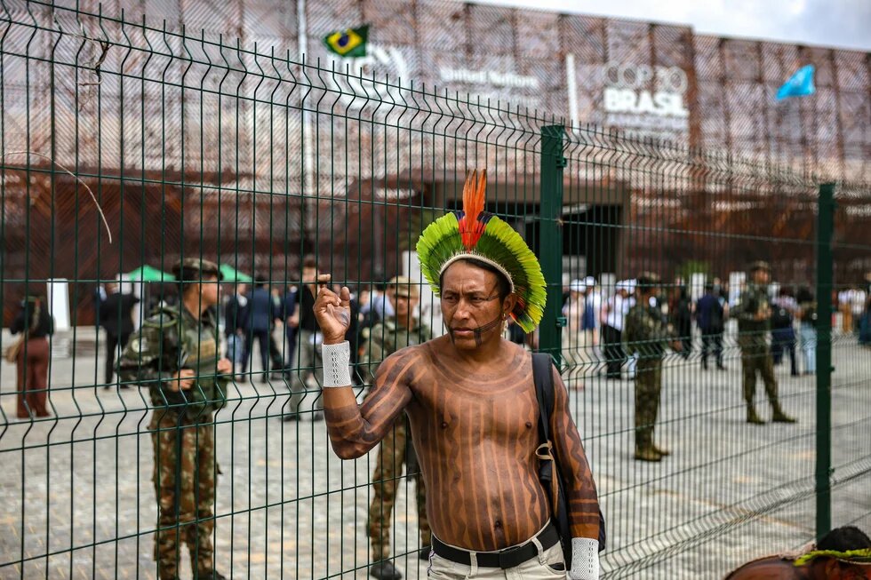 Person mit Federschmuck und Tätowierungen steht oberkörperfrei vor einem Zaun; im Hintergrund Militär und Menschen vor einem Gebäude mit der Aufschrift „COP30 BRASIL“.