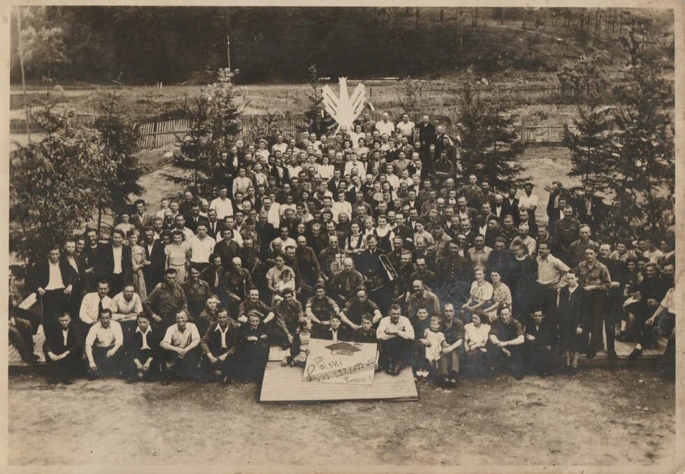 Große Menschengruppe sitzt und steht im Freien, vorne ein Schild mit Noten und Schrift, im Hintergrund Bäume und Zaun. Das Foto ist schwarz-weiß und wirkt alt. Es hat sichtbare Gebrauchsspuren und einen Sepia-Ton.