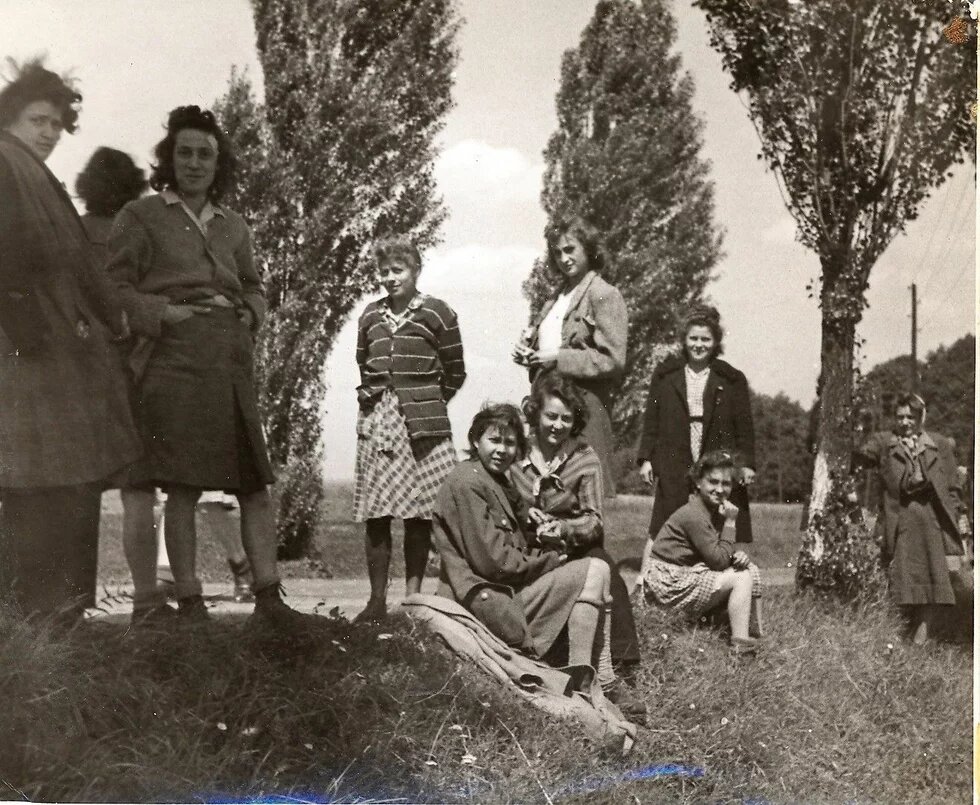 Gruppe von etwa zehn Personen steht oder sitzt auf Wiese mit Bäumen, teils lachend, Kleidung im Stil der 1940er Jahre.