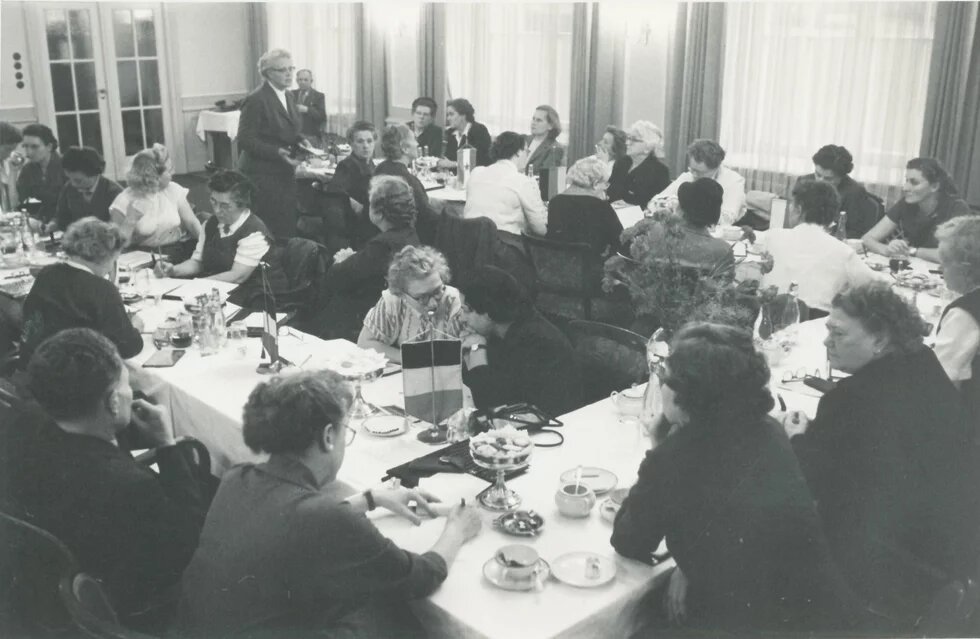Zahlreiche Personen sitzen an gedeckten Tischen, eine Frau steht sprechend am Rand, auf den Tischen stehen kleine Flaggen.