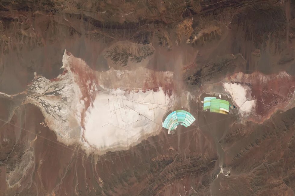 Bright salt flats with straight lines, turquoise-green basins for lithium mining on the right in a dry mountain landscape.