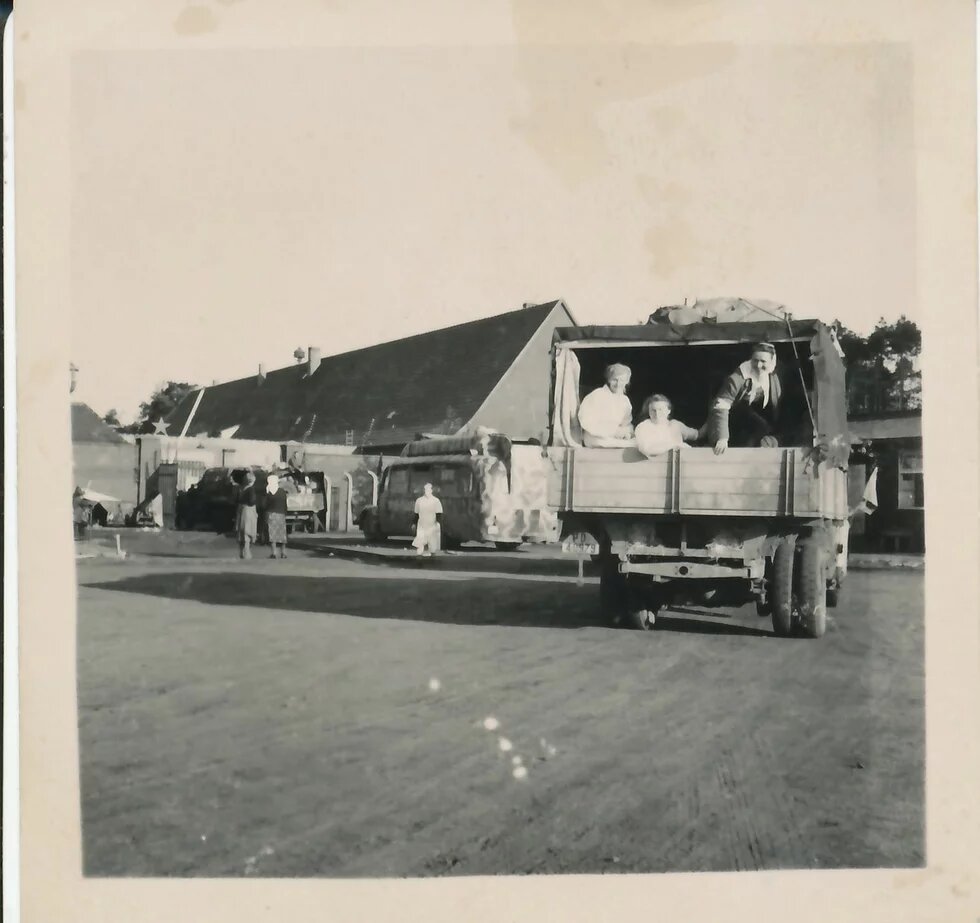 Drei Personen sitzen auf offener Ladefläche eines LKWs, weitere Menschen und Fahrzeuge vor Gebäuden im Hintergrund.