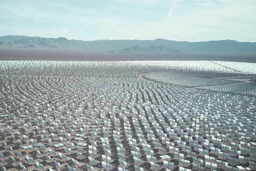Thousands of mirrors stand in rows in a desert landscape, with low mountains in the background under a lightly clouded sky.