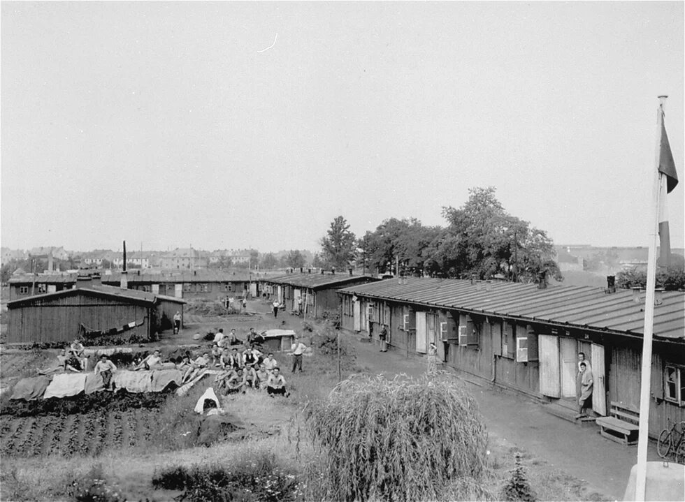 Mehrere Baracken am Weg, davor viele Menschen bei Gartenarbeit, Wäsche hängt, einzelne Personen stehen oder gehen zwischen den Gebäuden.