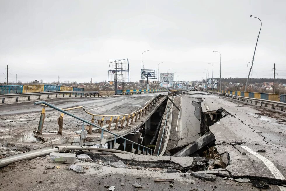 Destroyed road bridge with collapsed center section; guardrail bent, roadway severely damaged, city in the background.