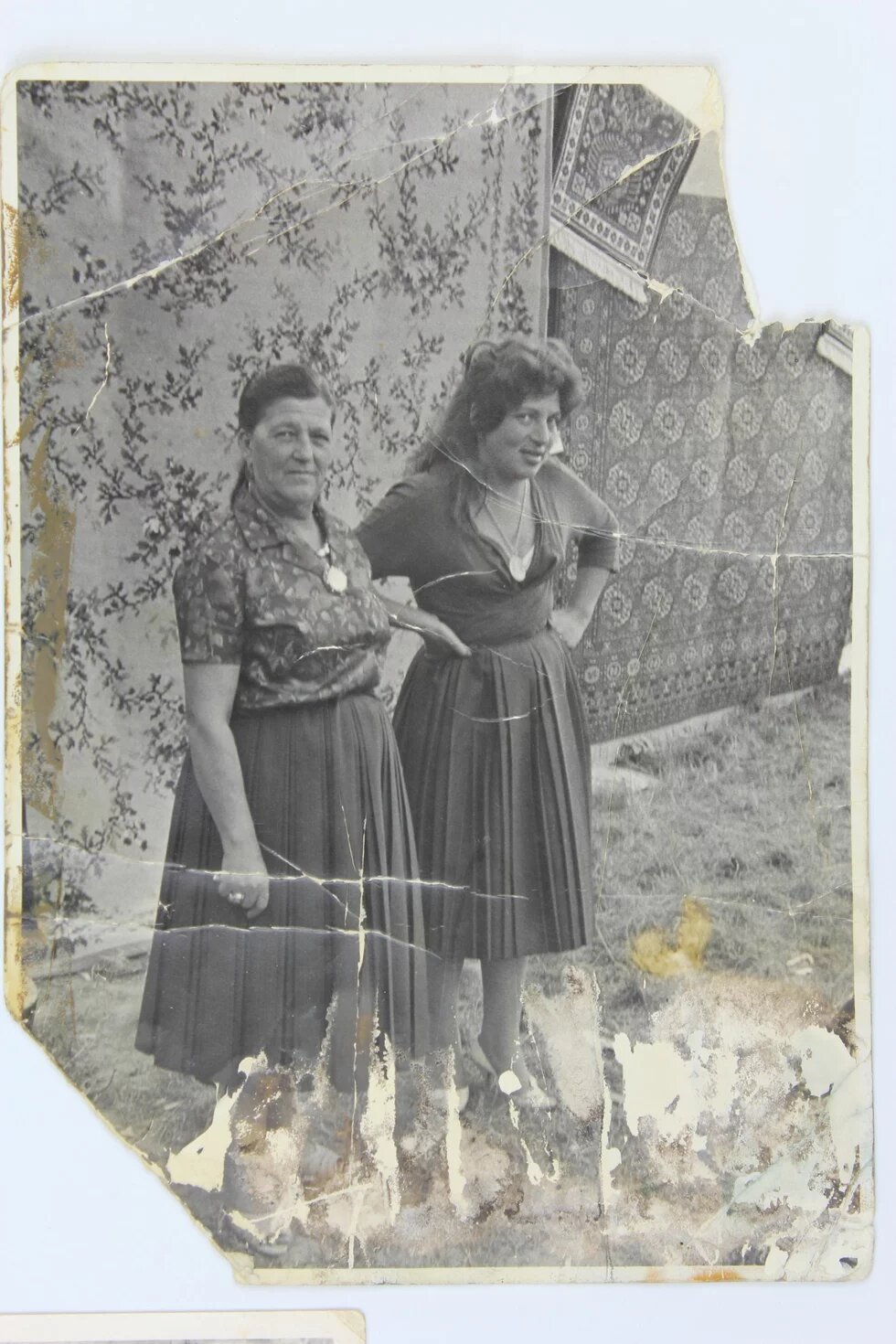 Zwei Personen in Röcken stehen nebeneinander im Freien vor gemusterten Teppichen, eine blickt zur Kamera, die andere zur Seite. Foto stark beschädigt.