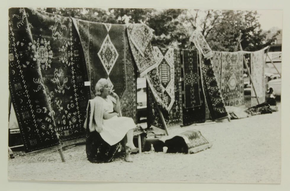 Person sitzt rauchend vor aufgestellten Teppichen im Freien, einige Teppiche liegen am Boden, Autos im Hintergrund.