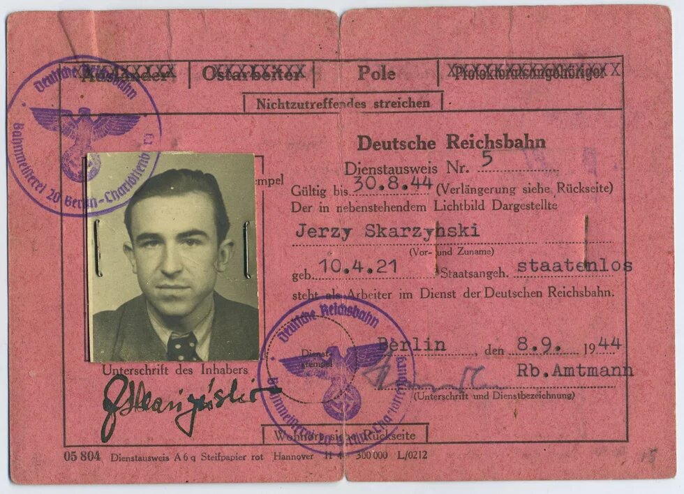 Rosa Dienstausweis der Deutschen Reichsbahn mit Lichtbild, Stempeln, Unterschriften und maschinenschriftlichen Angaben zur Person Jerzy Skarzynski,Porträtfoto eines jungen Mannes mit glatten, nach hinten gekämmten Haaren, Anzug und gepunkteter Krawatte.
