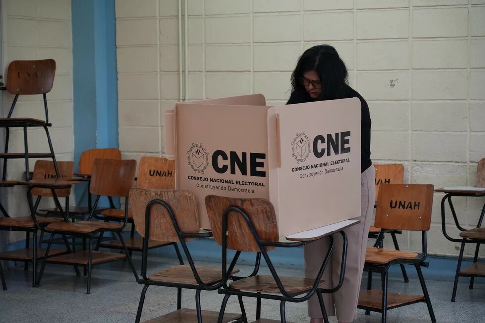 Eine Person steht an einer Wahlkabine mit der Aufschrift „CNE“ in einem leeren Raum mit gestapelten Schulstühlen.