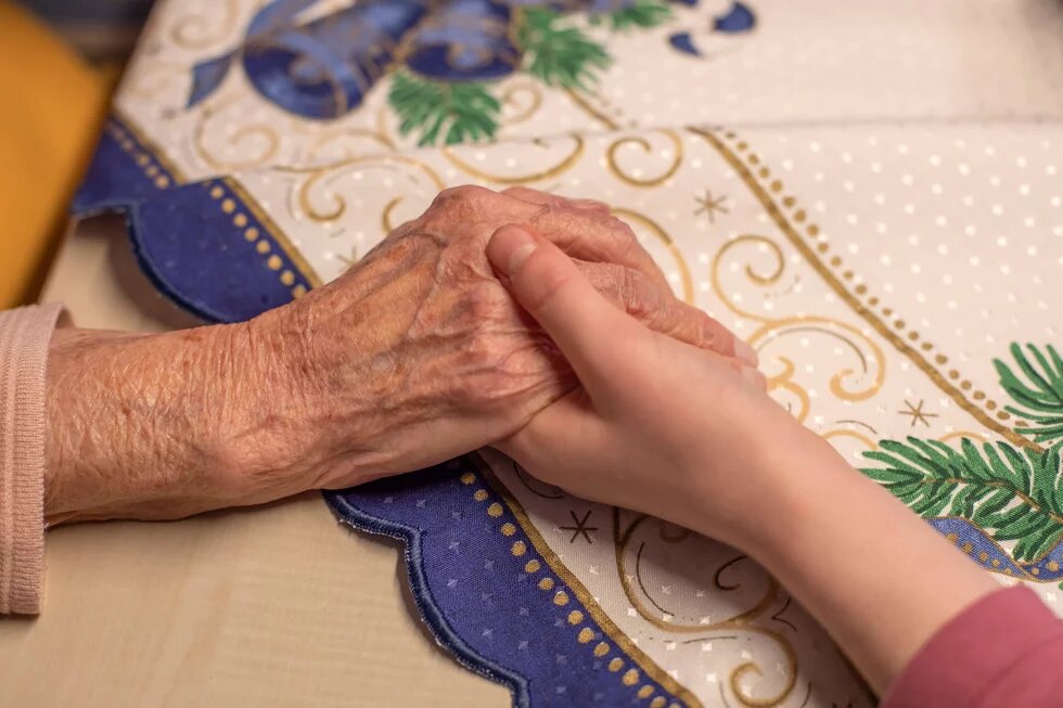 Eine junge Hand hält eine ältere Hand auf bestickter Tischdecke mit weihnachtlichem Muster.