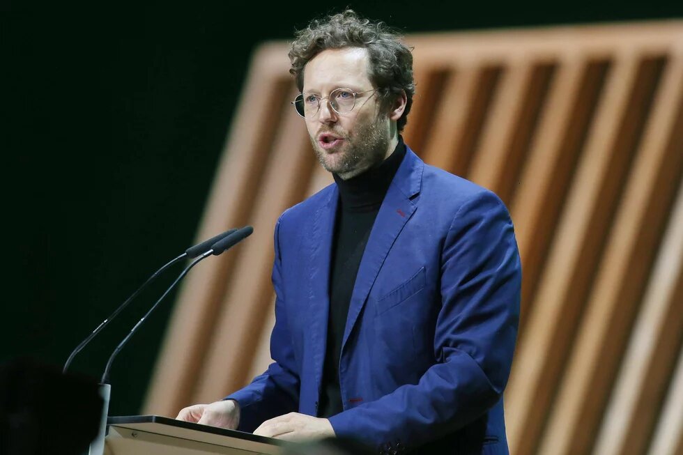 Ein Mann mit Brille und lockigem Haar spricht an einem Rednerpult mit zwei Mikrofonen, im Hintergrund schräge Holzlamellen.