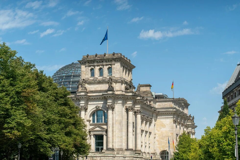 Reichstagsgebäude in Berlin mit Glaskuppel, deutscher und EU-Flagge, teils von Bäumen verdeckt.