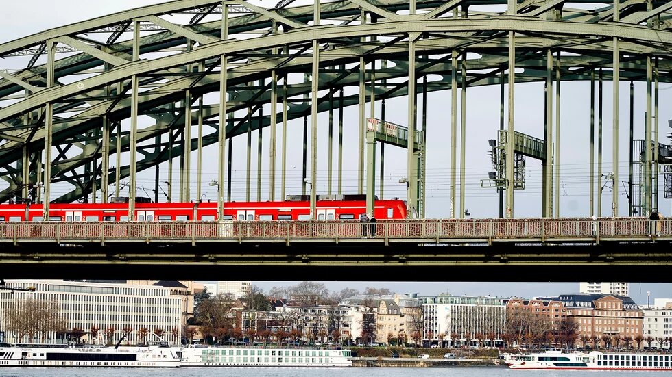 Rote Regionalbahn fährt über die Hohenzollernbrücke in Köln, darunter der Rhein mit zwei weißen Schiffen.
