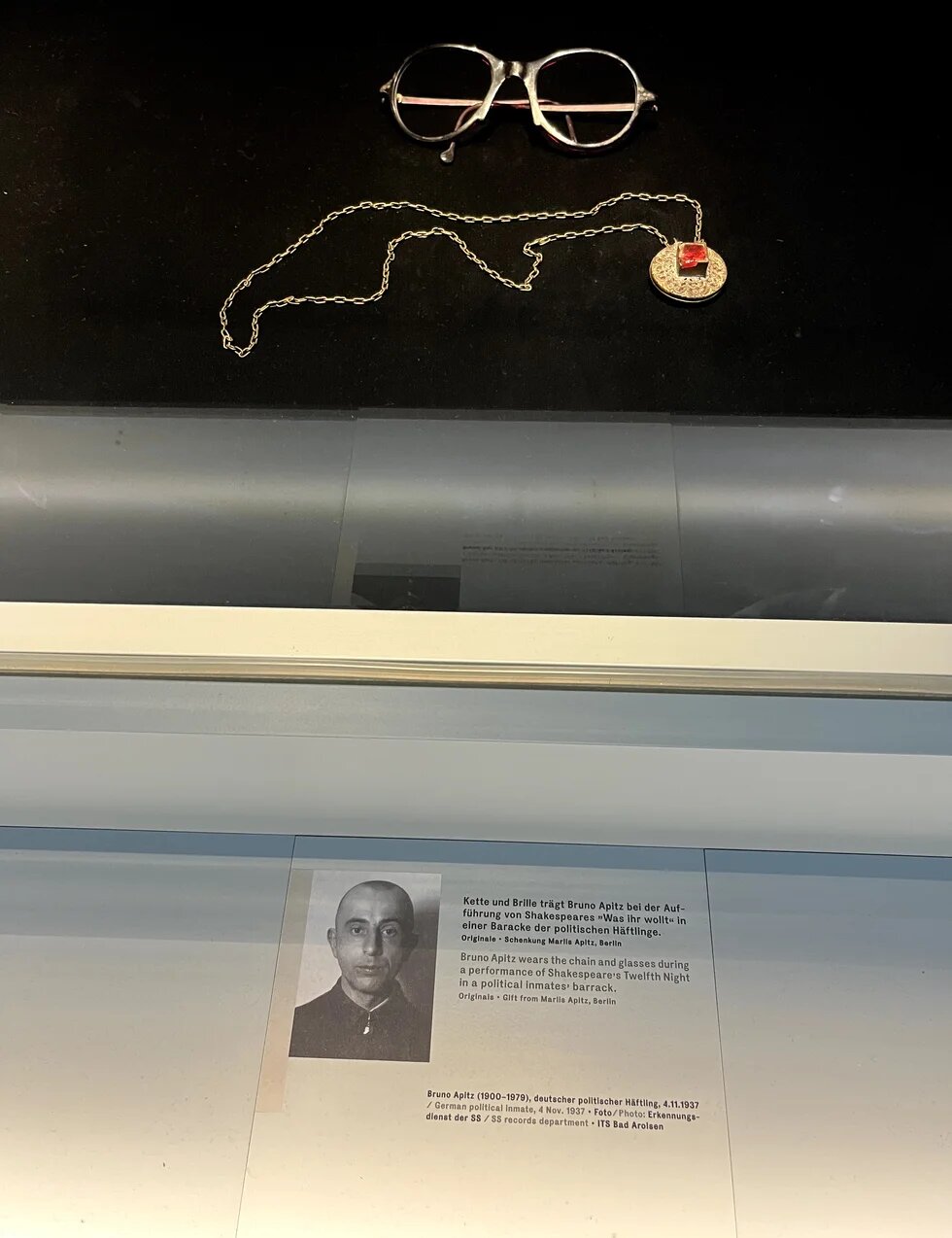 Brille und Kette mit Anhänger liegen in einer Vitrine; darunter ein Porträtfoto und Infotext zu Bruno Apitz, politischer Häftling.