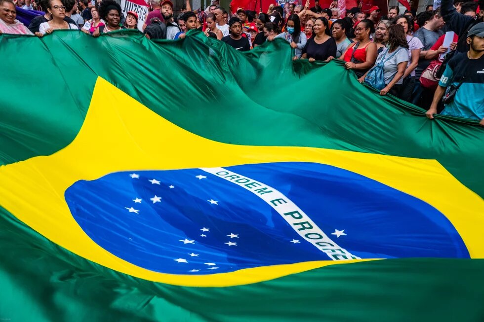 Eine große Menschenmenge hält gemeinsam eine riesige brasilianische Flagge hoch, die sich über den Vordergrund des Bildes spannt.