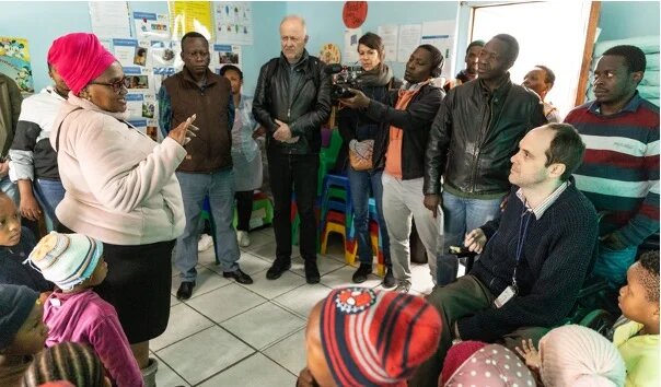 Eine Frau spricht vor einer Gruppe Erwachsener und Kinder in einem Raum mit bunten Stühlen und Infopostern an der Wand.