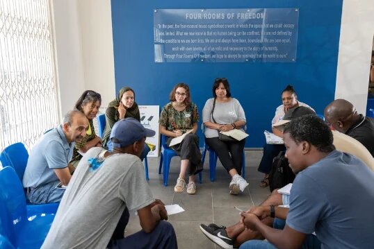 Two groups of people are sitting opposite each other on blue chairs and talking in a room with an information board on the wall.