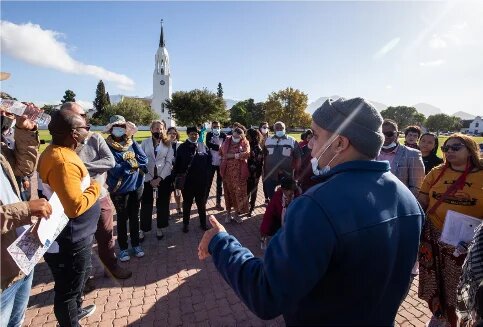 A large group stands in a circle in a square, one person is speaking, and a white church with a tower can be seen in the background.