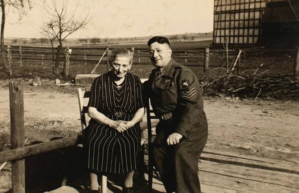 Zwei Personen sitzen draußen nebeneinander, eine ältere Person in Kleid, eine in Uniform; im Hintergrund Zaun und Felder.