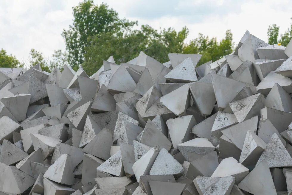 Viele graue Betonelemente in Pyramidenform liegen dicht gestapelt vor grünem Baumbewuchs.