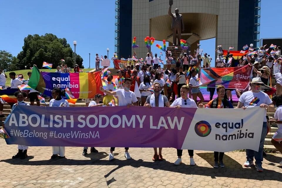 Many people stand in front of a monument on a staircase with rainbow flags and a banner from the organization Equal Namibia that reads “ABOLISH SODOMY LAW.”