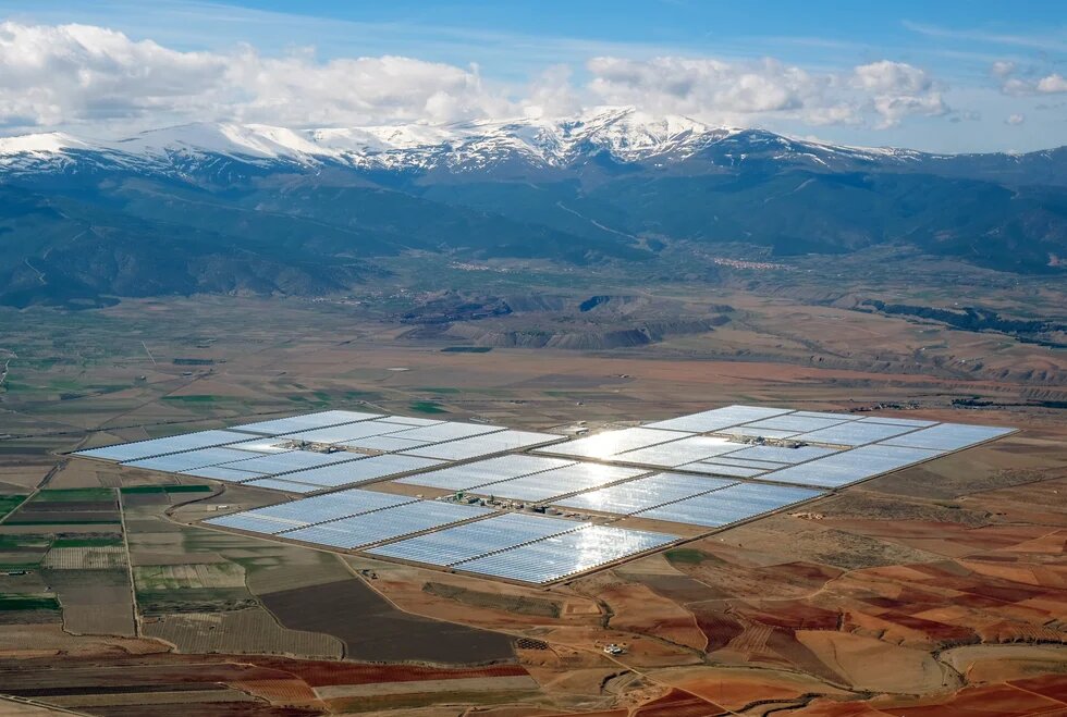 A large solar power plant made up of reflective panels spreads across a flat, arid landscape, with mountains visible in the distance under a partly cloudy sky.