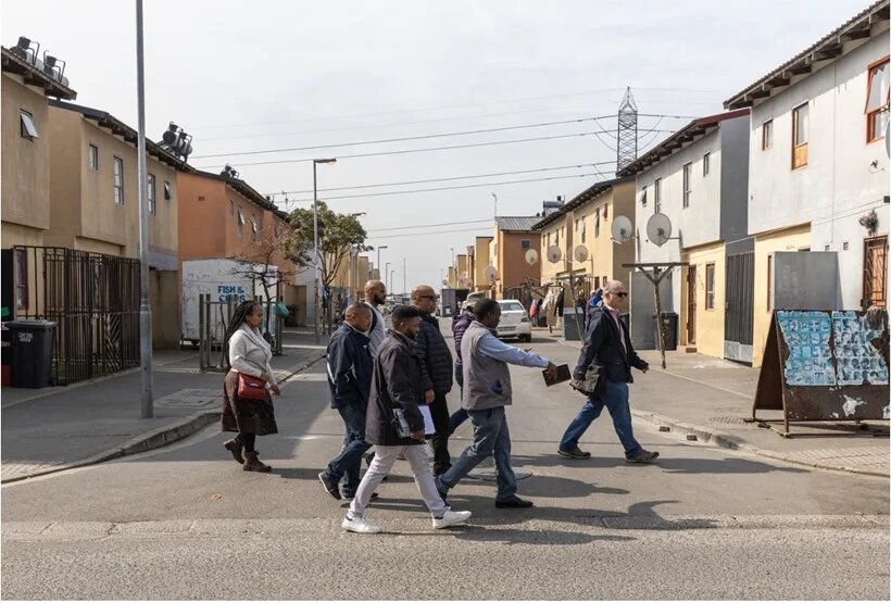 Das Foto zeigt eine Gruppe von Menschen, die eine Straße in einem südafrikanischen Township überquert, was ein typisches städtisches Wohngebiet am Rande von Städten oder Gemeinden darstellt. 