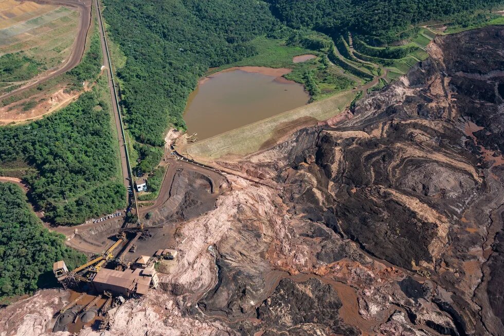 Aerial view shows mine, dam, brown basin, mud flowing into valley.