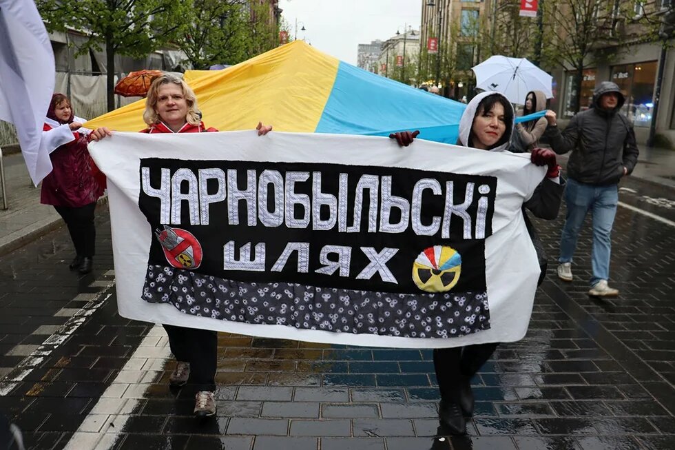 Zwei Frauen halten bei Regen auf einer Straße ein Banner mit kyrillischer Aufschrift, während hinter ihnen Menschen unter einem großen blau-gelben Tuch und mit Regenschirmen gehen.